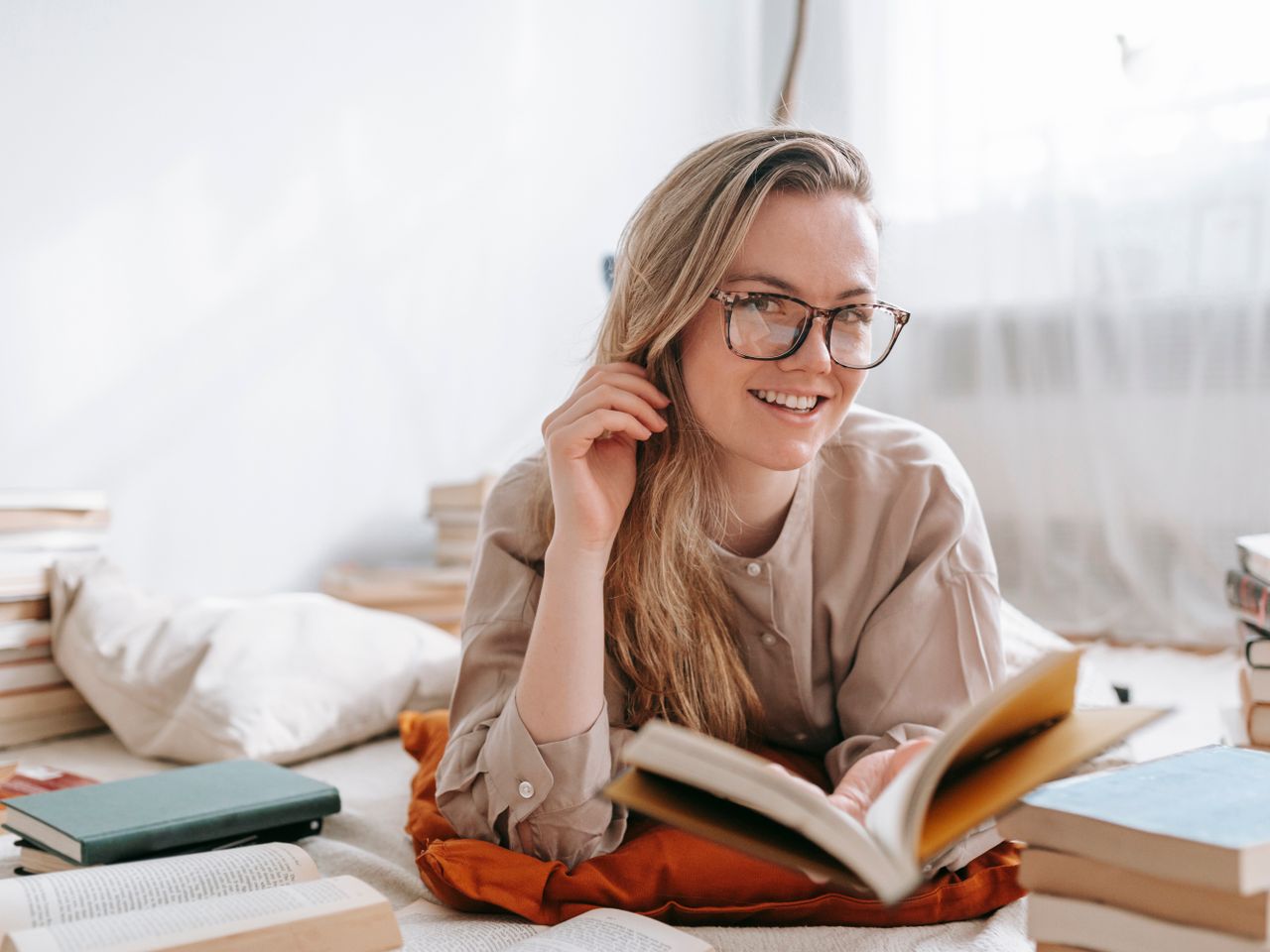 A person smiling while reading a book at home, surrounded by books.